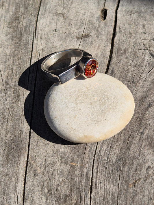 A Sterling Silver and Champagne Cubic Zirconia Ring with a sleek modern design, featuring a round, warmed champagne-colored gemstone set on a narrow silver band, placed on a textured wooden surface.
