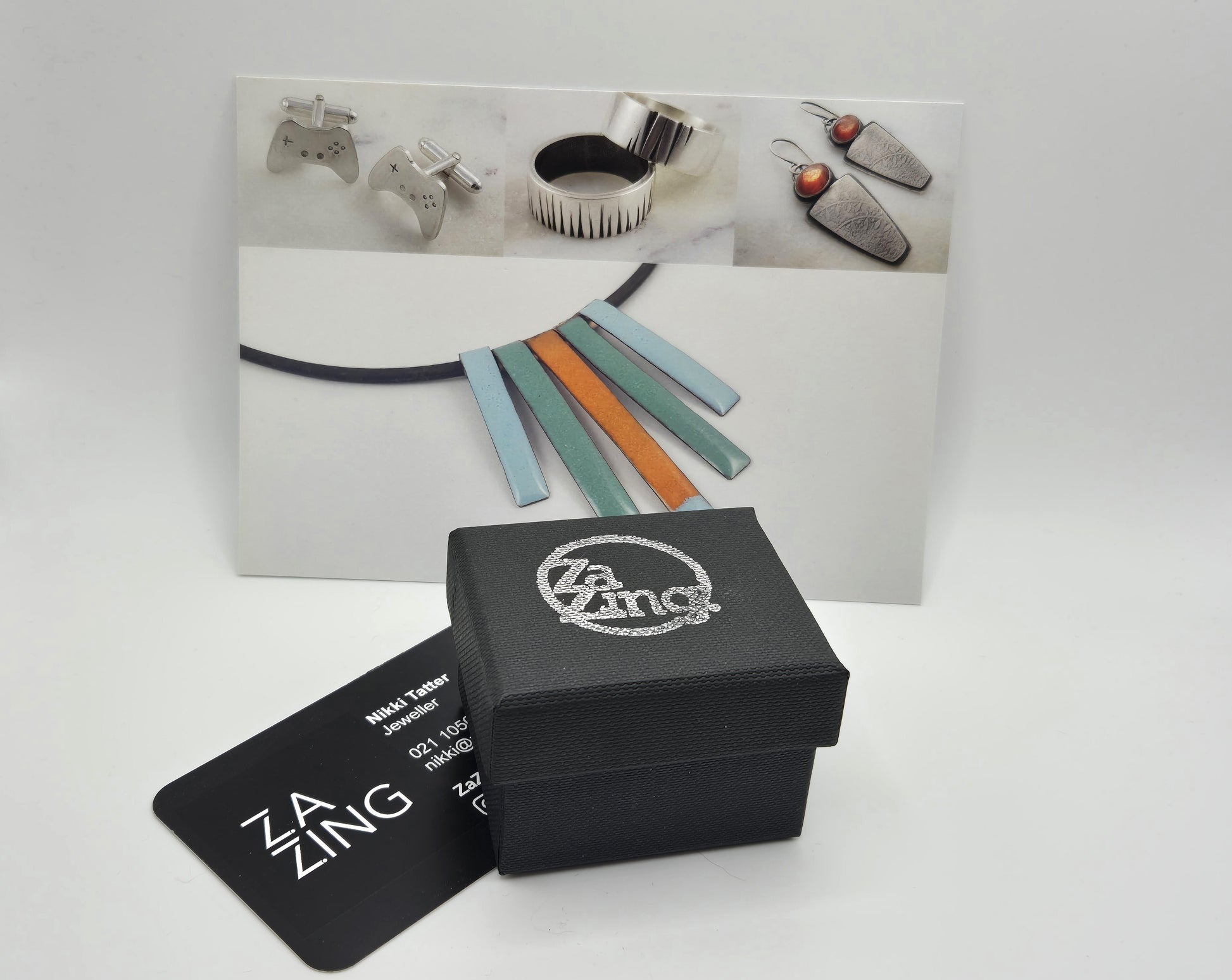 ZaZing Packaging including black ring box and business card.  In the background is a postcard with ZaZing jewellery on it