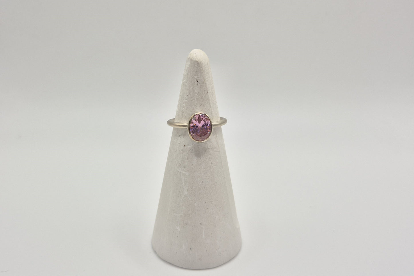 A slim sterling silver band ring with a bezel-set pink cubic zirconia stone positioned at the top, displayed on a white cone-shaped pedestal.