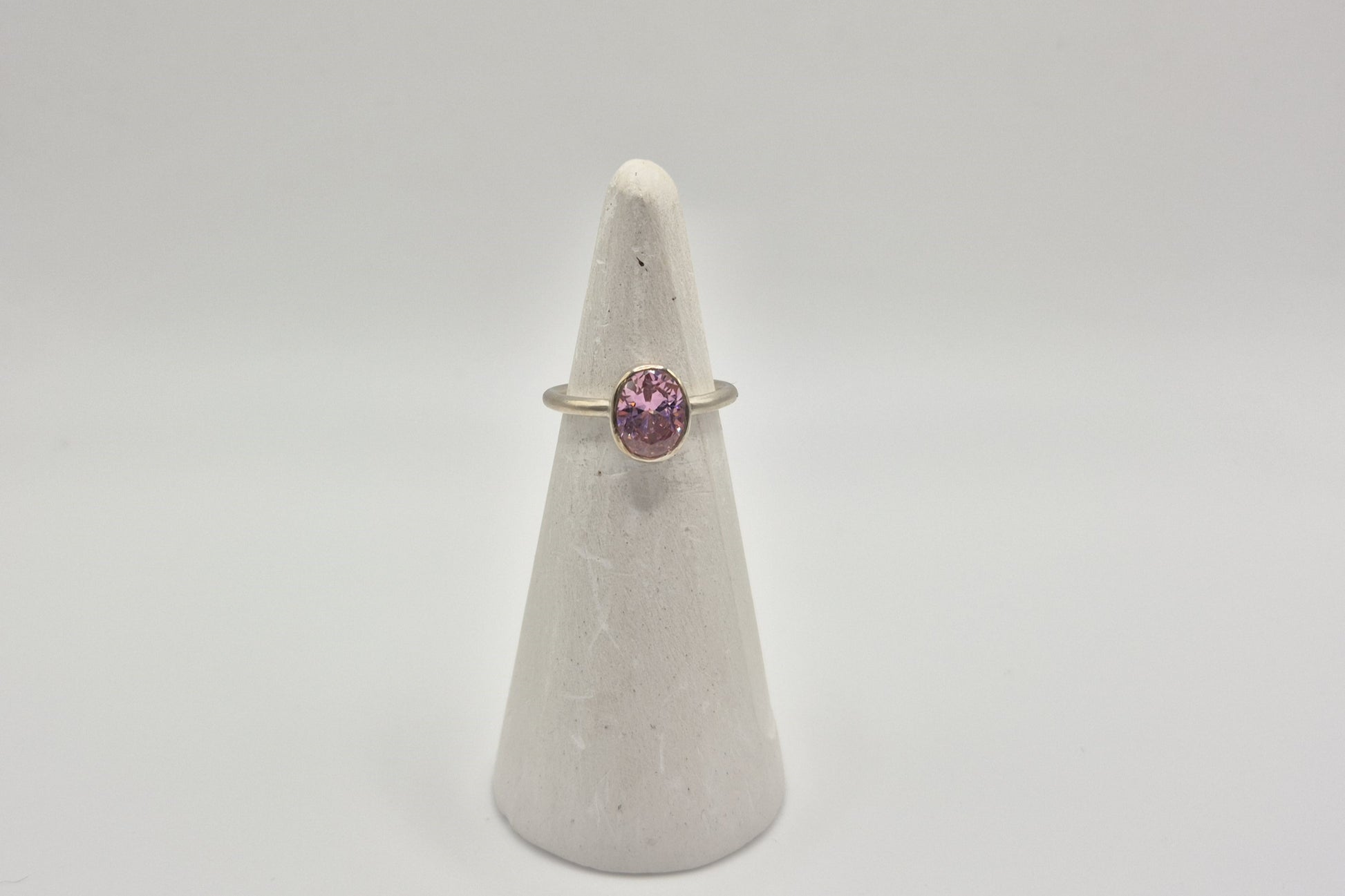 A slim sterling silver band ring with a bezel-set pink cubic zirconia stone positioned at the top, displayed on a white cone-shaped pedestal.
